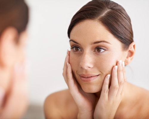 A gorgeous young woman looking at her reflection in the mirror with her hands on her face. Laser Hair Removal in New Jersey